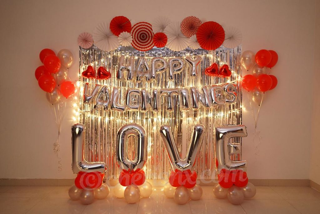 Happy Valentine’s Love Decor Surprise featuring frill curtain on the wall along with red and white rosettes, and white and red balloons. 
