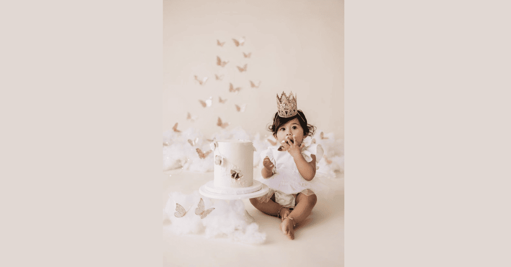 Captivating dreamy ambiance: Child in white gown and crown enjoying a cake during her 1st birthday photoshoot.