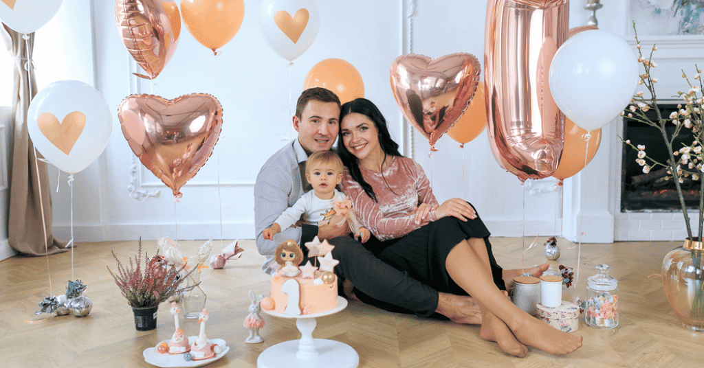  Child seated on a lap of parents with birthday cake, surrounded by heart-shaped foil and pastel balloons, capturing love and togetherness for a perfect birthday indoor photoshoot.