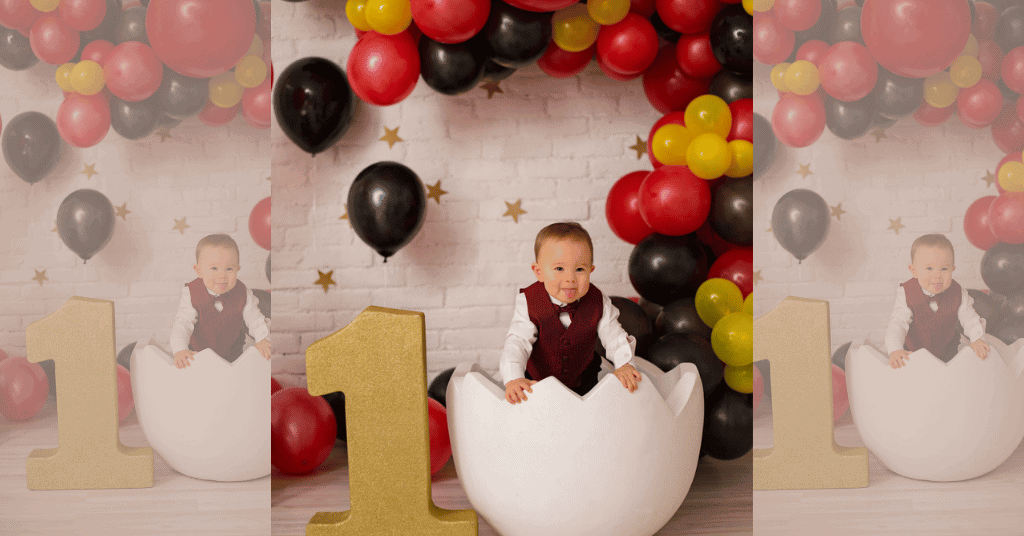  1st birthday photoshoot idea where a child is inside a half-cracked egg seat with vibrant Mickey Mouse-themed balloons, symbolizing the emergence of a milestone.