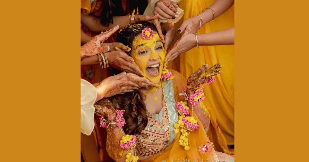 The bride to be laughing while there are hands applying Haldi on her face 